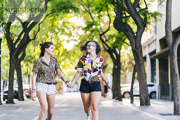 Glückliches lesbisches Paar hält sich an den Händen und läuft auf dem Fußweg in der Stadt an einem sonnigen Tag