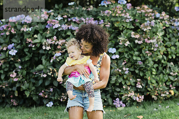 Mittlere erwachsene Frau  die ein kleines Mädchen trägt  während sie vor einer blühenden Pflanze im Park steht