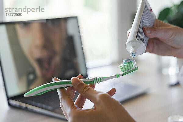 Hände einer Zahnärztin  die Paste auf eine Zahnbürste aufträgt  die durch einen Videoanruf an einen Patienten gezeigt wird