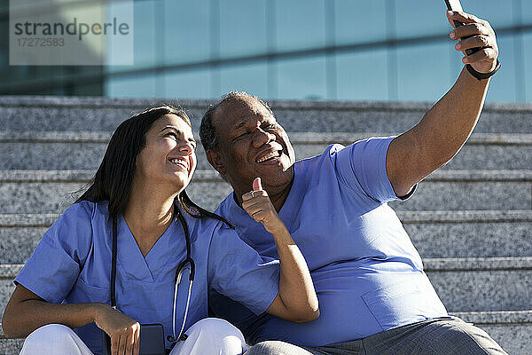 Lächelnde Ärzte  die ein Selfie machen  während sie auf der Treppe vor dem Krankenhaus sitzen