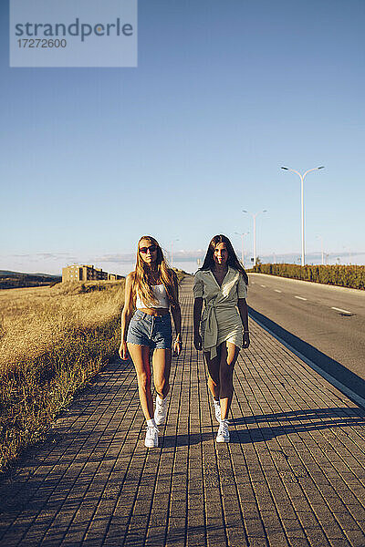 Junge Freundinnen gehen auf dem Bürgersteig gegen den Himmel an einem sonnigen Tag