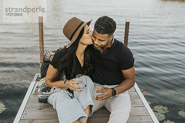 Frau mit Weinglas küsst Mann auf dem Pier sitzend