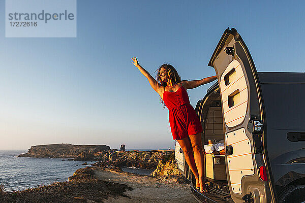 Lächelnde Frau mit erhobener Hand  die an der Tür eines Wohnmobils am Strand hängt