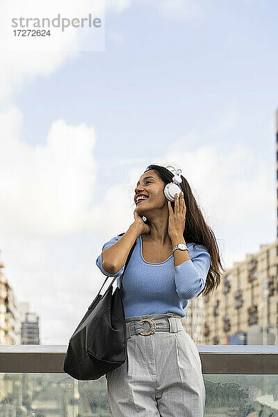 Glückliche Geschäftsfrau  die Musik über Kopfhörer hört  während sie sich an das Geländer gegen den Himmel lehnt