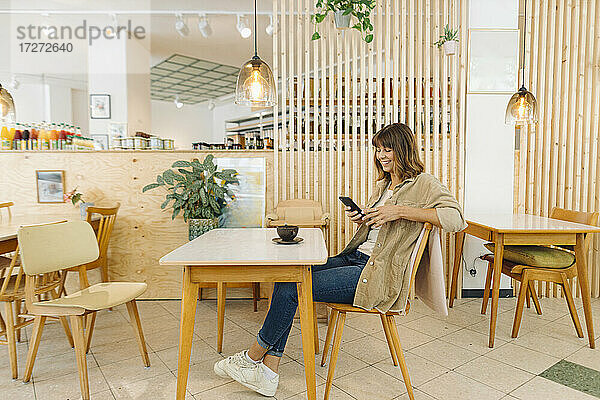 Lächelnde Geschäftsfrau  die in einem Café sitzend eine Textnachricht auf ihrem Smartphone schreibt