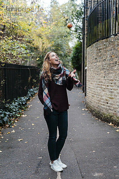 Lächelnde Frau  die einen Apfel fängt  während sie auf einem Fußweg im Herbst steht