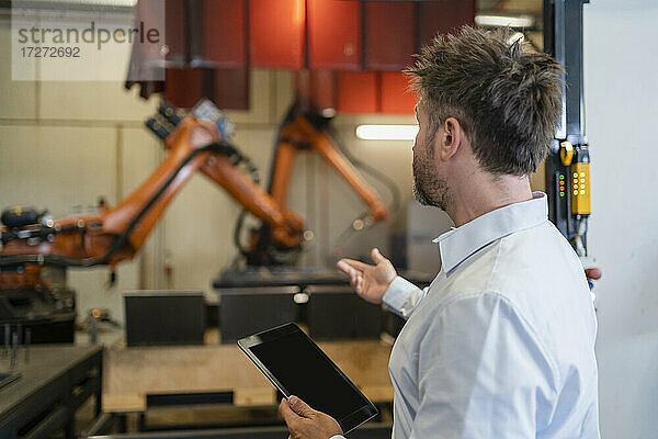 Älterer Geschäftsmann mit digitalem Tablet  der in Richtung einer Roboterarm-Maschine gestikuliert  während er in einer Fabrik steht