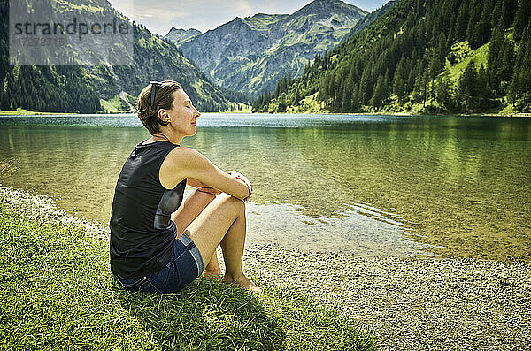 Entspannte Frau sitzt mit geschlossenen Augen am Seeufer an einem sonnigen Tag