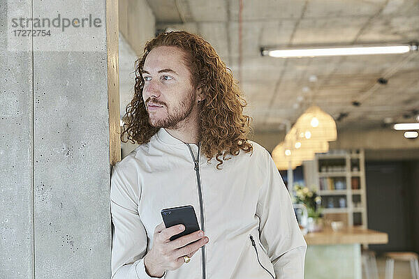 Hipster-Mann schaut weg  während er sein Smartphone an eine Betonsäule zu Hause lehnt