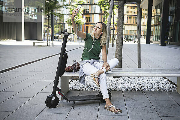 Lächelnd Mitte erwachsene Frau hören Musik und nehmen selfie beim Sitzen auf Bank in der Stadt