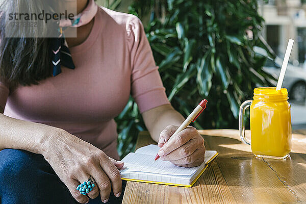 Reife Frau schreibt in Tagebuch  während sie mit frischem Saft am Tisch in einer Bar sitzt