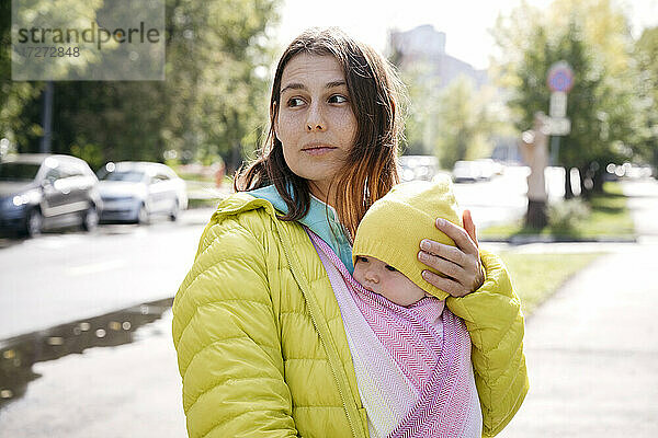 Mutter hält ihr in eine Decke eingewickeltes Baby  während sie an einem sonnigen Tag auf der Straße steht