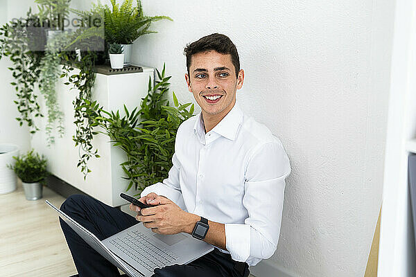 Lächelnder gutaussehender Unternehmer sitzt mit Laptop und Smartphone vor einer weißen Wand in einem kreativen Büro