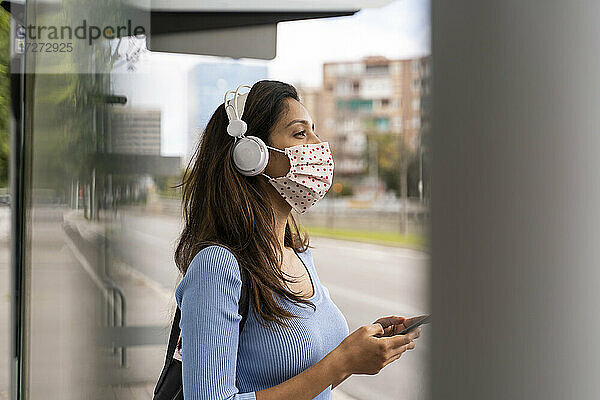 Nachdenkliche Frau mit Gesichtsmaske  die Kopfhörer trägt und ihr Smartphone am Busbahnhof während COVID-19 benutzt