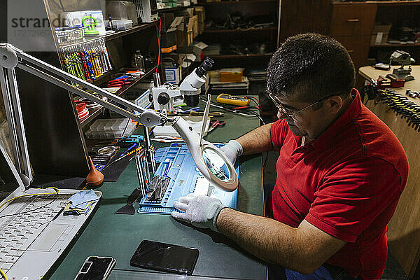 Arbeiter  der ein kaputtes Smartphone durch ein Vergrößerungsglas in einer Reparaturwerkstatt betrachtet