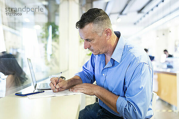Selbstbewusster männlicher Berufstätiger  der mit seinem Laptop in einem Café sitzt und auf Papier schreibt