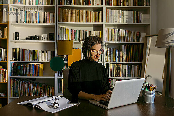 Lächelnde Geschäftsfrau  die an einem Laptop arbeitet und zu Hause am Bücherregal sitzt