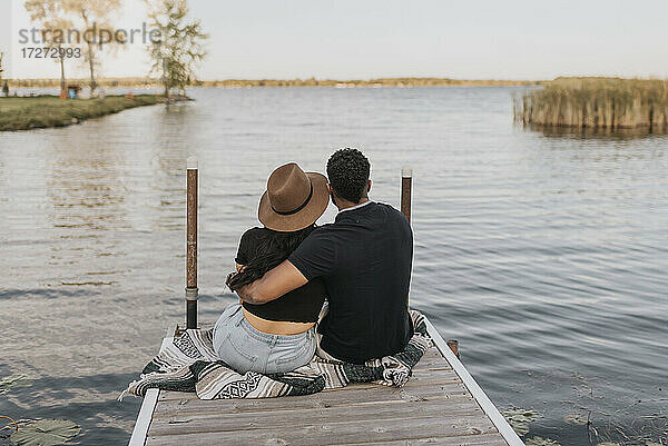 Paar mit Arm um bewundernde Ansicht beim Sitzen auf Pier gegen See