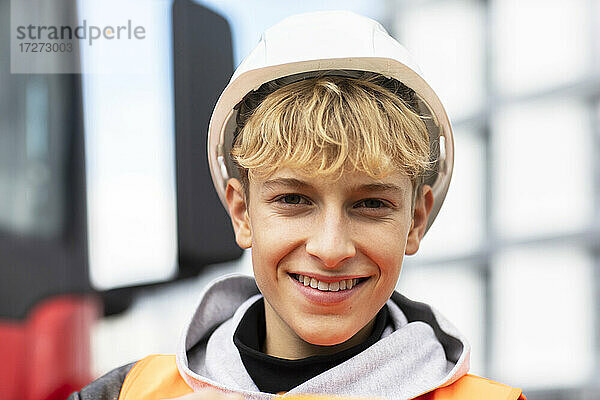 Lächelnder blonder männlicher Jugendlicher mit Schutzhelm auf einer Baustelle