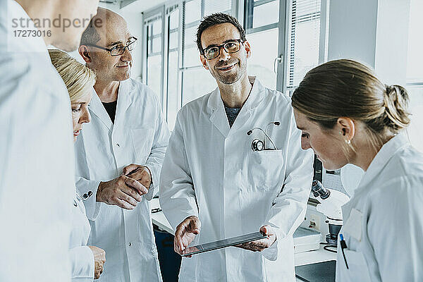 Lächelnder Wissenschaftler  der ein digitales Tablet in der Hand hält  während er neben seinen Kollegen im Labor steht