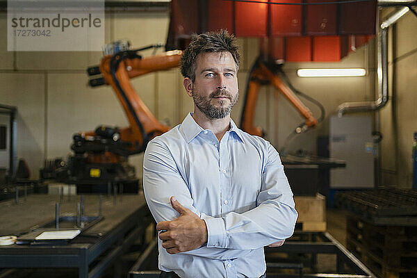 Selbstbewusster Geschäftsmann  der mit verschränkten Armen vor einer Roboterarm-Maschine in einer Fabrik steht