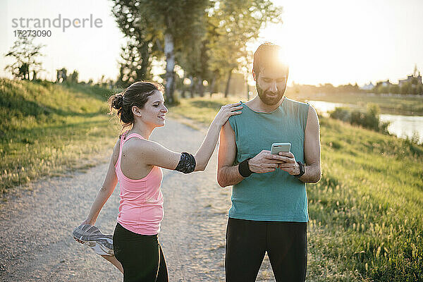 Männlicher Sportler  der sein Smartphone benutzt  während eine Sportlerin mit hochgelegtem Bein im Park bei Sonnenuntergang steht