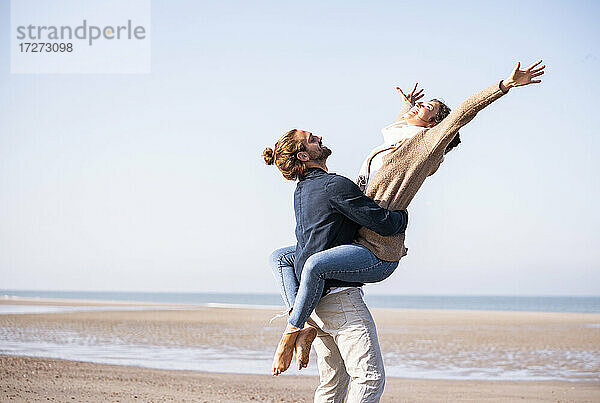 Junger Mann trägt glückliche Freundin mit ausgestreckten Armen am Strand gegen klaren Himmel an einem sonnigen Tag