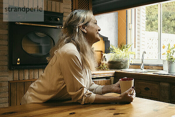 Lächelnde Frau  die wegschaut  während sie in der Küche zu Hause Kaffee trinkt