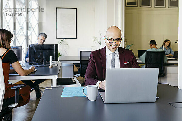 Lächelnder glatzköpfiger Geschäftsmann arbeitet am Laptop am Schreibtisch im Büro