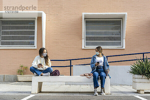 Freundinnen  die auf einer Betonbank sitzend eine Gesichtsmaske tragen und sich sozial distanzieren