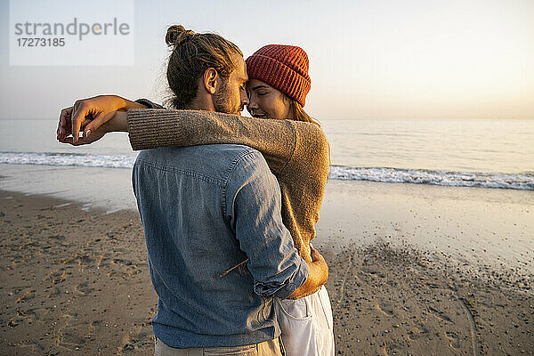 Junges romantisches Paar steht von Angesicht zu Angesicht am Strand gegen den klaren Himmel bei Sonnenuntergang