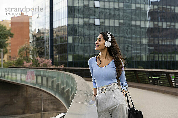 Lächelnde Geschäftsfrau hört Musik über Kopfhörer  während sie auf einem Steg vor einem modernen Gebäude steht