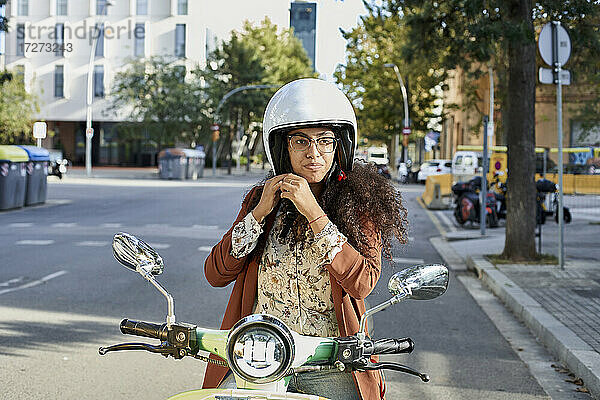 Junge Frau befestigt ihren Helm  während sie auf einem Motorroller in der Stadt sitzt