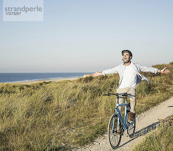 Unbekümmerter Mann mit ausgestreckten Armen auf dem Fahrrad am Strand gegen den klaren Himmel