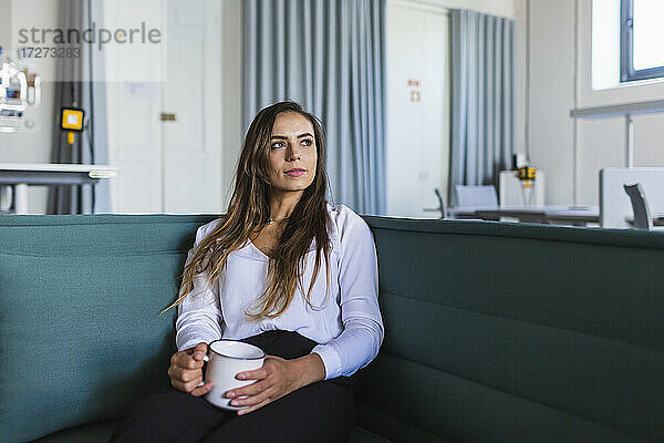 Nachdenkliche Geschäftsfrau mit Kaffeetasse auf dem Sofa im Büro
