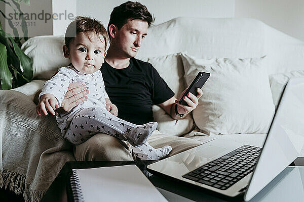 Vater benutzt sein Smartphone  während er seinen Sohn auf dem Sofa zu Hause hält