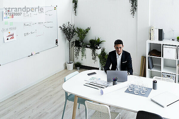 Lächelnder Geschäftsmann  der einen Laptop benutzt  während er am Schreibtisch sitzt und eine Strategie im Büro plant