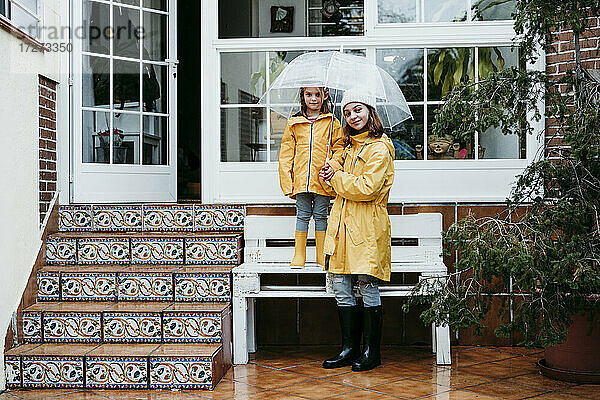 Lächelnde Schwester  die einen Regenschirm hält  während ihre Schwester auf einer Bank vor dem Haus steht
