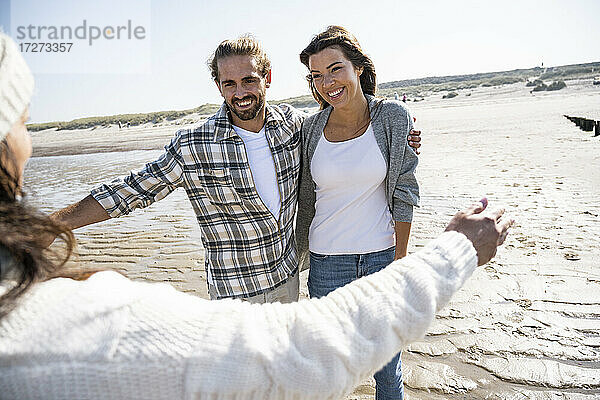 Reife Frau mit erhobenen Armen  die ein am Strand stehendes Paar umarmt