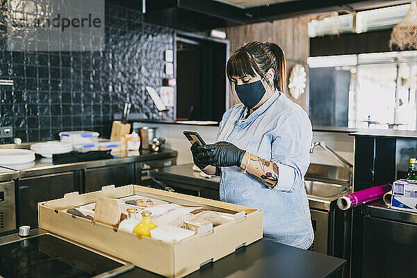 Weiblicher Koch  der ein Smartphone benutzt  während er an der Küchentheke eines Restaurants steht  in dem das Coronavirus grassiert.