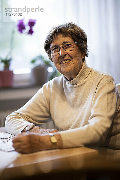 Lächelnde faltige Frau mit Notfallknopf am Handgelenk  die zu Hause am Tisch sitzt