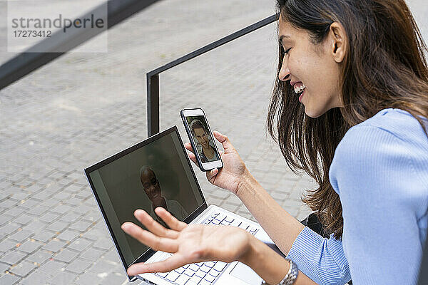 Glückliche Geschäftsfrau  die während der COVID-19 im Freien sitzend Videogespräche über Laptop und Smartphone führt