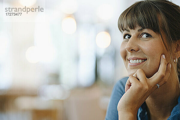 Lächelnde Unternehmerin  die mit der Hand am Kinn in einem Café sitzt und wegschaut