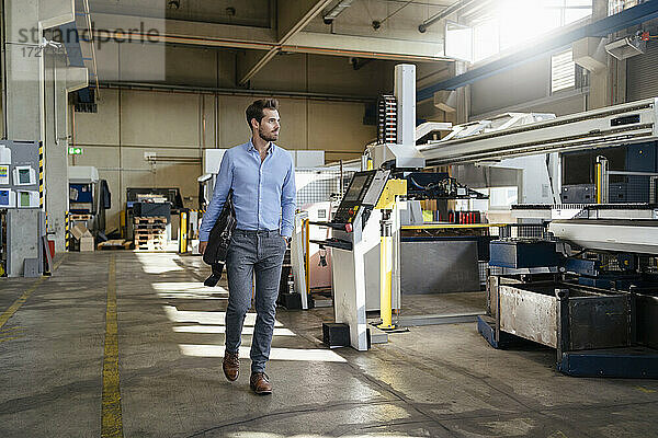 Junger Geschäftsmann hält Aktentasche beim Spaziergang in einer Fabrik