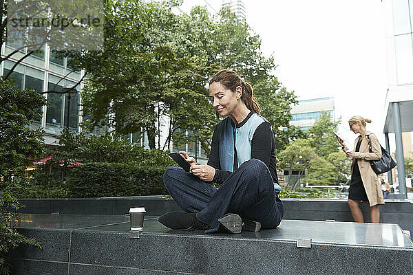 Geschäftsfrau  die ein Mobiltelefon benutzt  während sie auf einer Bank sitzt  während ein Kollege im Hintergrund im Büropark steht
