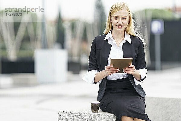 Junge Geschäftsfrau benutzt ein digitales Tablet  während sie auf einer Bank vor einem Gebäude sitzt