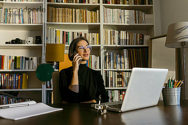 Geschäftsfrau  die mit einem Smartphone spricht und wegschaut  während sie zu Hause am Laptop sitzt und auf ein Bücherregal schaut