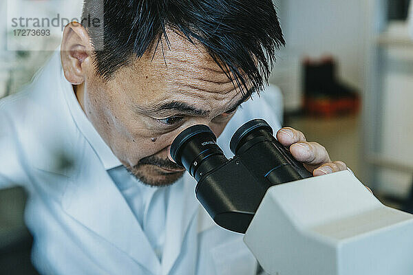 Männlicher Wissenschaftler schaut durch ein Mikroskop  während ein Mann im Hintergrund im Labor steht