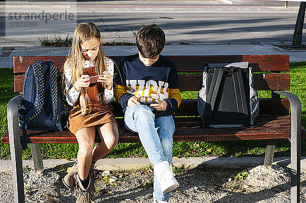 Geschwisterkind  das sein Smartphone benutzt  während es auf einer Bank in einem öffentlichen Park bei Sonnenschein sitzt