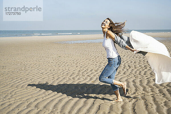 Unbeschwerte junge Frau  die an einem sonnigen Tag am Strand läuft und eine Decke hält